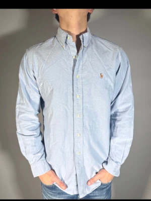 Ljusblå skjorta från Ralph Lauren - Klassisk ljusblå skjorta från Ralph Lauren med button-down krage och broderad logga på bröstet. Skjortan har långa ärmar och knäppning framtill. Perfekt för en clean och stilren look.