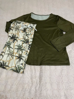 Grön pyjamas med palmbyxor - Mysig pyjamas med långärmad, olivgrön tröja och matchande byxor i vitt med palmmönster och kedjedetaljer. Perfekt för chillkvällar hemma. Mjukt och bekvämt material som känns skönt mot huden.