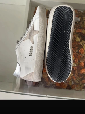 Golden Goose - Snygga vita sneakers från Golden Goose med den ikoniska stjärnan på sidan och grå detaljer. Skorna har en slitstark gummisula, grå skosnören och är tillverkade i läder med mockadetaljer. Lite skadad sula, skriv privat för närbild.