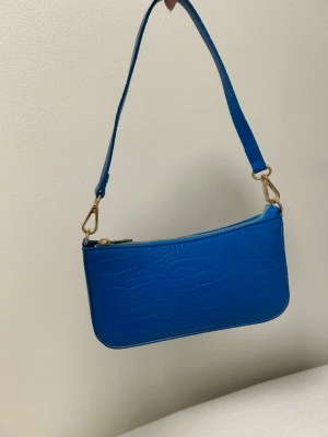 Blå axelväska i krokopräglat material - Säljer en trendig blå handväska med krokopräglat mönster och guldfärgade detaljer. Väskan har en avtagbar axelrem och stängs med dragkedja. Perfekt för dig som vill ha en färgklick till din outfit.