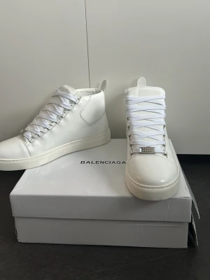 Balenciaga arena high  - Stilrena vita high top sneakers från Balenciaga i slätt läder. Klassisk siluett med vita skosnören och metallisk detalj framtill på snörningen. Kommer med originalkartong och har en clean look som passar till det mesta. Använda endast en gång boxen är tyvärr sönder.