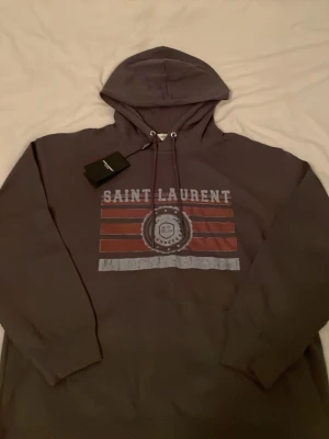 Mörkgrå Saint Laurent hoodie - Snygg mörkgrå hoodie från Saint Laurent med tryck i vitt och rött framtill. Klassisk huva med dragsko och ribbade muddar vid ärmar och nederkant. Hoodien har en casual vibe och är tillverkad i mjukt material för extra komfort.