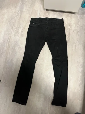 Svarta stretchjeans från BOSS - Säljer ett par svarta jeans från BOSS med smal passform och stretchigt material för extra komfort. Jeansen har klassisk femficksdesign, bälteshällor och knappgylf. Perfekta för dig som gillar stilrena och tidlösa plagg.