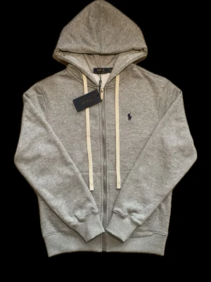 Grå zip hoodie från Polo Ralph Lauren - Snygg grå hoodie med dragkedja från Polo Ralph Lauren. Klassisk design med vit dragsko i huvan och mörkblå logga broderad på bröstet. Tillverkad i mjuk bomullsblandning och har ribbade muddar vid ärmar och midja. Perfekt för en avslappnad streetwear-look.