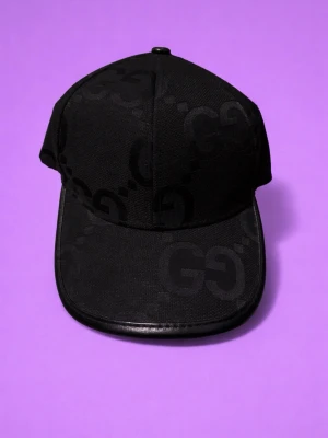 Svart keps från Gucci med GG-mönster - Snygg svart keps från Gucci med det klassiska GG-monogrammet ton-i-ton över hela kepsen. Skärmen har en stilren kant i svart läder och insidan är fodrad med band där det står Gucci. Perfekt accessoar för en trendig streetwear-look.