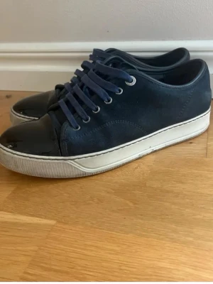 Mörkblå sneakers från Lanvin - Snygga mörkblå sneakers från Lanvin med vita sulor och lackade tåpartier. Skorna har blå skosnören och en klassisk låg profil. Materialmix av mocka och lack ger en exklusiv känsla. Perfekta för dig som gillar stilrena och trendiga sneakers.