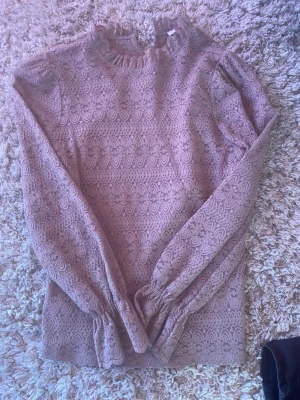 Brun/beige spetsblus med volang - Supersöt brun/begie spetsblus med långärmad design och volangdetaljer vid krage och ärmslut. Blusen har ett vackert blommigt spetsmönster och är lätt transparent. Perfekt för dig som gillar romantisk och trendig stil. Dålig kamera därför syns färgen dåligt 