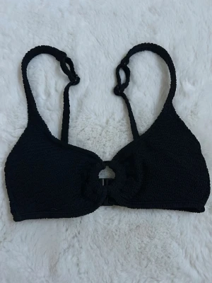 Svart bikiniöverdel med ring - Stilren svart bikiniöverdel med struktur och justerbara axelband. Modellen har en rund uringning och dekorativ öppning framtill samt spänne i ryggen. Perfekt för strandhäng eller pooldagar. Använd max 3 gånger