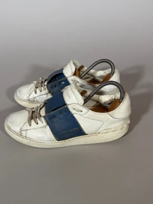 Valentino vita och blå sneakers - Snygga sneakers från Valentino i vitt läder med breda blåa detaljer på sidorna. Klassisk låg modell med vita snören och diskret logga på plösen. Skorna har en chunky sula och stilren design som passar till många olika outfits.