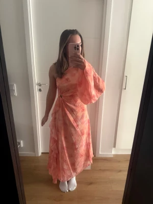 Oneshoulder långklänning i rosa toner - Säljer en flowy oneshoulder långklänning i ljusrosa och orange toner med ett subtilt blommigt mönster. Klänningen har en voluminös ärm och markerad midja med drapering och ringdetalj. Perfekt för dig som vill sticka ut med en unik och feminin look.