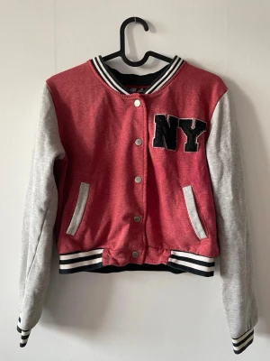 Röd collegejacka med NY-tryck - Röd collegejacka från B- Soul med grå ärmar och svartvita ribbade muddar. Tryckknappar framtill och broderad NY-logga på bröstet. Två fickor och klassisk varsity-stil. Perfekt för en sportig och trendig look.