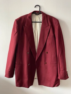 Vinröd kavaj - Stilren vinröd kavaj med breda slag. Kavajen har guldfärgade knappar och två framfickor. Fodrad insida i ljus färg. 