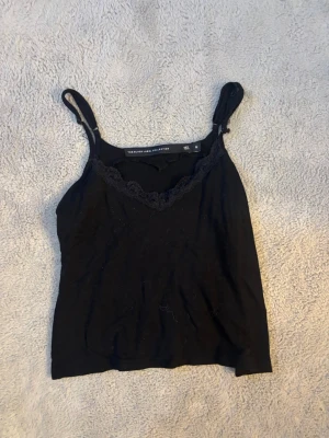 Svart spetsdetaljerad linnetopp - Snygg svart linnetopp från The Black Label Collection med tunna axelband och spetsdetalj längs v-ringningen. Perfekt att styla lager på lager eller bära som den är. Mjuk och skön känsla i materialet. Lite nopprig men jätte mysig!