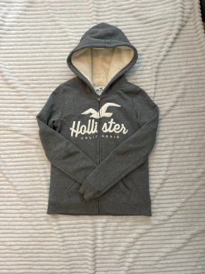 Hollister fodrad grå zip up hoodie med dragkedja - Mysig grå hoodie från Hollister med vit logga och text på bröstet.                                                                    Storlek XS!                                                             Hoodien har dragkedja framtill, fodrad i hela hoodien. Perfekt för chill dagar och enkel att matcha med jeans.