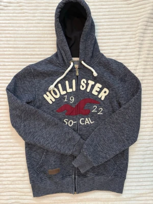 Hollister zip up hoodie - Mörkblå grå zip up hoodie från Hollister med dragkedja, vit brodyrtext och röd logga på bröstet.     Storlek S!                                                                      Stor huva med vita snören. Två fickor framtill och ribbade muddar vid ärmslut och nederkant. Perfekt för en chill och avslappnad stil.