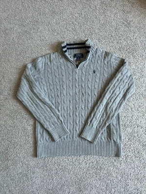 Grå kabelstickad tröja Polo Ralph Lauren - Snygg grå kabelstickad tröja från Polo Ralph Lauren med halv dragkedja och ribbade muddar. Tröjan har en liten mörkblå logga broderad på bröstet och en randig detalj på insidan av kragen. Perfekt för lager på lager och chill stil.