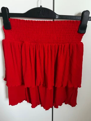 Röd volangkjol från Kappahl  - Säljer en röd kortkjol från kappahl med bred smock i midjan och dubbla volanglager. Kjolen har en luftig och flowy känsla, perfekt för varmare dagar. Materialet är mjukt och lätt, vilket gör den superbekväm att bära.