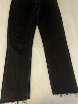 Svarta raka jeans med rå kant - Säljer ett par svarta jeans med rak passform och råa, fransiga benslut. Jeansen har en klassisk femficksmodell och är tillverkade i denim. Perfekta för en avslappnad och trendig look.