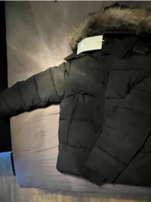 Svart dunjacka från Canada Goose - Säljer en svart dunjacka från Canada Goose med klassisk patch på ärmen och stor huva med fuskpäls. Jackan är tjockt vadderad och har en rak passform, perfekt för kalla vinterdagar. Stängs med dragkedja och tryckknappar.