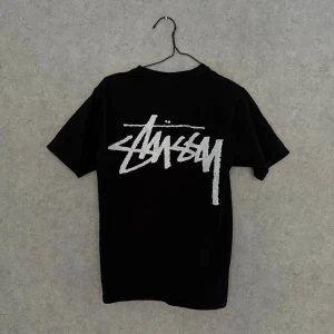 Stüssy/Our Legacy Tee - Svart t-shirt från Stüssy x Our Legacy med stort vitt logotryck på ryggen och en liten yin-yang-symbol på bröstet. Klassisk passform och rund hals. Perfekt för dig som gillar streetwear och vill ha en enkel men snygg tee.