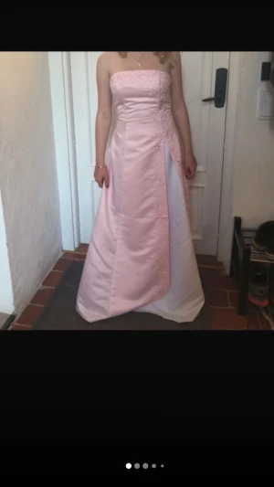 Ljusrosa axelbandslös balklänning - Vintage axelbandslös långklänning i ljusrosa och vitt med broderade detaljer och omlott-design. Klänningen har en klassisk A-linjeform och är perfekt för bal eller student. Materialet ser ut att vara satin eller polyester för en glansig finish. Klänningen har en liten fläck längst ned vid linningen, men annars perfekt skick! 