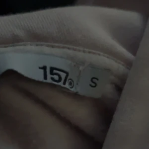 Ljusrosa hoodie från 157 - Mysig ljusrosa hoodie från 157 med klassisk känguruficka och huva. Tröjan har ribbade muddar vid ärmslut och nederkant, perfekt för en avslappnad och chill stil. Passar till jeans eller mjukisbyxor för en enkel vardagslook.