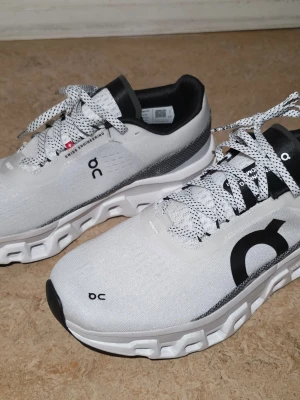 Vita On Running sneakers med svart detalj - Snygga vita sneakers från On Running med svart logga och detaljer. Skorna har en unik sula med vågformad design och vita, svarta samt grå inslag. Ovandelen är i mesh-liknande material för bra andningsförmåga och skorna har vita snören med svarta prickar. SNABB AFFÄR 500KR