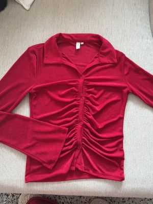 Röd rynkad blus från Nelly - Trendig röd blus från Nelly med långa ärmar och snygg rynkning framtill. Blusen har klassisk krage, knappar längs hela framsidan och är gjord i ett mjukt, stretchigt material som sitter skönt på kroppen. Perfekt för dig som vill sticka ut lite extra.