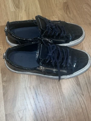 Guiseppe Zanotti Skor - Säljer ett par svarta sneakers med glansig krokodilpräglad yta, svarta skosnören och dragkedjor på sidorna. Skorna har vit sula och coola metalldetaljer vid hälen. Strl 43. Fraktar samma dag och bra skick p skorna