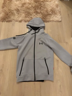 Grå hoodie från Under Armour - Snygg grå hoodie från Under Armour med dragkedja framtill och svarta detaljer. Hoodien har två fickor med dragkedja, justerbar huva med snören och logga på bröstet samt ärmen. Perfekt för en sportig och avslappnad stil. Aldrig använd helt ny 