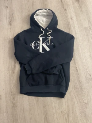 Svart hoodie från Calvin Klein - Snygg svart hoodie från Calvin Klein med stor vit CK-logga och text på bröstet. Hoodien har vit dragsko och fodrad huva i vitt tyg. Klassisk känguruficka framtill och ribbade muddar vid ärmar och nederkant. Perfekt för en avslappnad och trendig look.