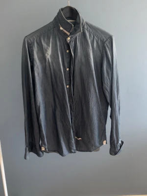 Mörkblå skjorta med knappar - Snygg mörkblå skjorta i bomull med klassisk krage och knäppning framtill. Skjortan har långa ärmar och en avslappnad passform, perfekt för en stilren look. Inga synliga mönster eller detaljer förutom knappar.