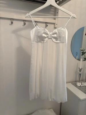 Vit klänning med rosett framtill - Sött vit klänning ny med tunna axelband och en stor dekorativ rosett framtill. Överdelen är i glansigt satinliknande material och kjoldelen är mjukt och lätt tyg. Smockad rygg för skön passform. 