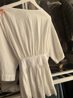 Vit omlottopp med knytband - Säljer en vit omlottopp med knytband i midjan och trekvartsärm. Toppen har en strukturerad yta och markerad midja som ger en snygg siluett. Perfekt till jeans eller kjol för en fräsch look.