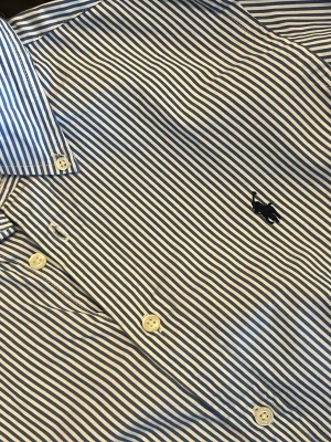 Blårandig skjorta från Ralph Lauren - Klassisk skjorta från Ralph Lauren med blå och vita ränder, broderad logga på bröstet och vita knappar framtill. Skjortan har en tidlös krage och är perfekt för dig som gillar preppy stil. Storleken är barn Xl men skulle säga att den passar S. 