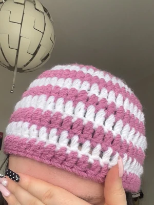Virkad rosa och vit randig mössa - Säljer en handvirkad mössa med breda ränder i rosa och vitt. Mössan har en klassisk form och är gjord i tjockt garn som ger en chunky och trendig look. Perfekt accessoar för dig som vill sticka ut med en färgglad och unik stil.