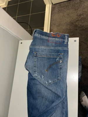 Blå slitna jeans med detaljer - Säljer ett par blå jeans med slitningar och trasiga detaljer framtill. Jeansen har klassisk femficksmodell och en avslappnad passform. Materialet är denim och de har en cool, använd look som passar perfekt till en streetwear-stil. Passar max 180-185 i längd och sitter som 32