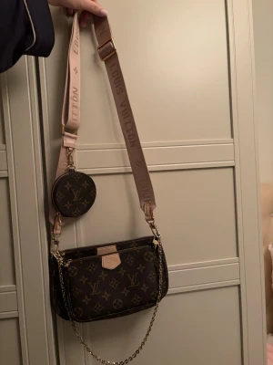  axelväska - Snygg axelväska i klassiskt brunt monogrammönster. Väskan har bred beige axelrem med logga, guldfärgade detaljer och en liten rund coin purse som hänger på remmen. Materialet är canvas med läderdetaljer.