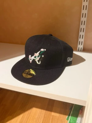 New Era 59Fifty keps - Säljer en helt oanvänd svart New Era 59Fifty keps med broderad logga framtill i vitt, grönt och rosa samt '1995 World Series' patch på sidan. Klassisk flat brim och broderade ventilationshål. Perfekt för dig som gillar streetwear och sportig stil.