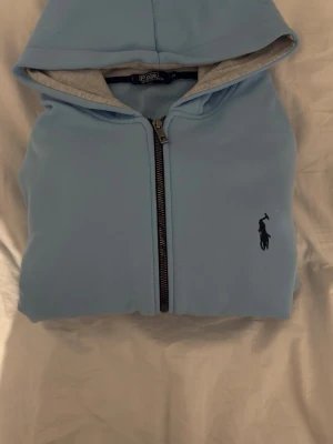 Ljusblå hoodie från Polo Ralph Lauren - Snygg ljusblå hoodie från Polo Ralph Lauren med dragkedja och mörkblå broderad logga på bröstet. Utsidan är i mjuk bomull och insidan av huvan är grå. Perfekt för en chill och stilren look.