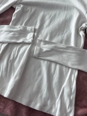 Vit ribbad långärmad topp - Snygg vit långärmad polo topp med ribbad struktur. Toppen har en enkel och clean design som passar till allt. Materialet känns mjukt och stretchigt, perfekt för lager på lager eller att bära som den är.