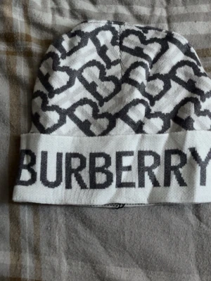 Vit och grå Burberry mössa - Säljer en snygg vit mössa från Burberry med grått hjärtmönster och stor logga framtill. Mössan är ribbstickad och har en uppvikt kant. Perfekt accessoar för att lyfta din outfit med en exklusiv touch.