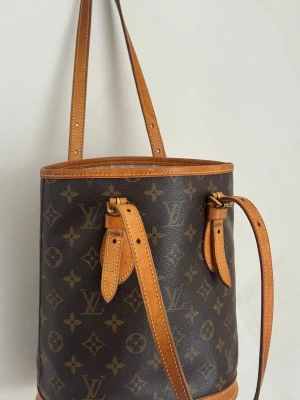 Louis Vuitton bucket bag - Säljer denna vackra och autentiska Louis Vuitton bucket bag i klassisk monogramcanvas i väldigt fint skick!  Den har en vacker patina på lädret vilket gör att den ser ännu lyxigare ut, samt små repor på detaljer och handtag, perfekt för älskare av vintage! Inuti väskan finns små slitage och flagning av fodret, väldigt ren och fin.  Storlek 26x23x16