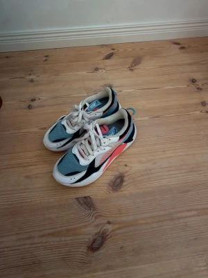 Puma sneakers i blått och rosa - Snygga sneakers från Puma med vit sula, blå mesh på ovandelen, svarta och vita detaljer samt en rosa sidopanel. Skorna har vita skosnören och en sportig design med låg profil. Perfekta för dig som gillar färg och streetstyle.
