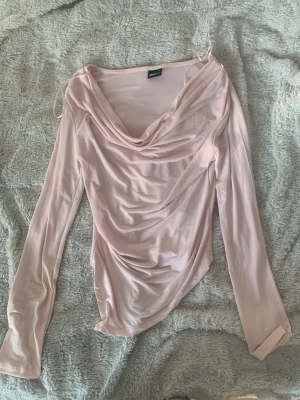 Ljusrosa omlottopp med drapering - Supersnygg ljusrosa volang topp från Gina Tricot med draperad framsida och omlottkänsla. Toppen har långa ärmar och en lös, lite glansig passform som ger en trendig look. Perfekt att styla med jeans eller kjol för en chic vibe. Helt nytt skick, har aldrig använts!