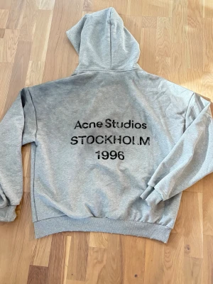 Grå hoodie Acne Studios Stockholm - Snygg grå hoodie från Acne Studios med tryck på ryggen där det står 'Acne Studios STOCKHOLM 1996'. Klassisk passform med huva och ribbade muddar. Perfekt för dig som gillar streetwear och enkel stil. Materialet är mjuk bomullsblandning.