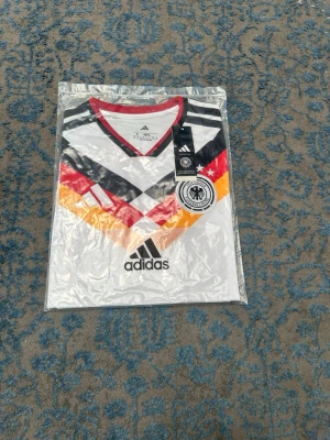 Tysklands retro fotbollströja Adidas - Vit retro fotbollströja från Adidas med Tysklands landslagsmärke. Tröjan har klassiska svarta, röda och gula mönster över bröstet, svart Adidas-logga och röd krage. Korta ärmar med detaljer i samma färger. Perfekt för fotbollsfans!