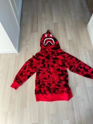 Röd BAPE Shark hoodie camo - Säljer en ikonisk röd BAPE Shark hoodie med camomönster och dragkedja hela vägen. Huvan har det klassiska hajansiktet med tänder och ögon. Tillverkad i mjuk bomullsblandning och har ribbade muddar vid ärmar och nederkant. Perfekt statement-plagg för streetwear.