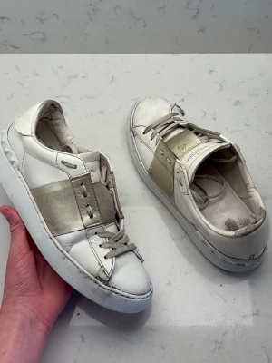 Valentino Garavani vita sneakers - Snygga vita sneakers från Valentino Garavani med bred guldfärgad rem över ovandelen och klassiska snörningar. Skorna har en låg profil och detaljerade nitar på hälen. Tillverkade i läder med gummisula och cool design som sticker ut.