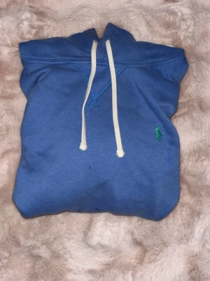 Blå hoodie från Polo Ralph Lauren - Snygg blå hoodie från Polo Ralph Lauren med klassisk känguruficka och vita snören.Storleken är S men den passar även XS. Liten grön logga broderad på bröstet. 
